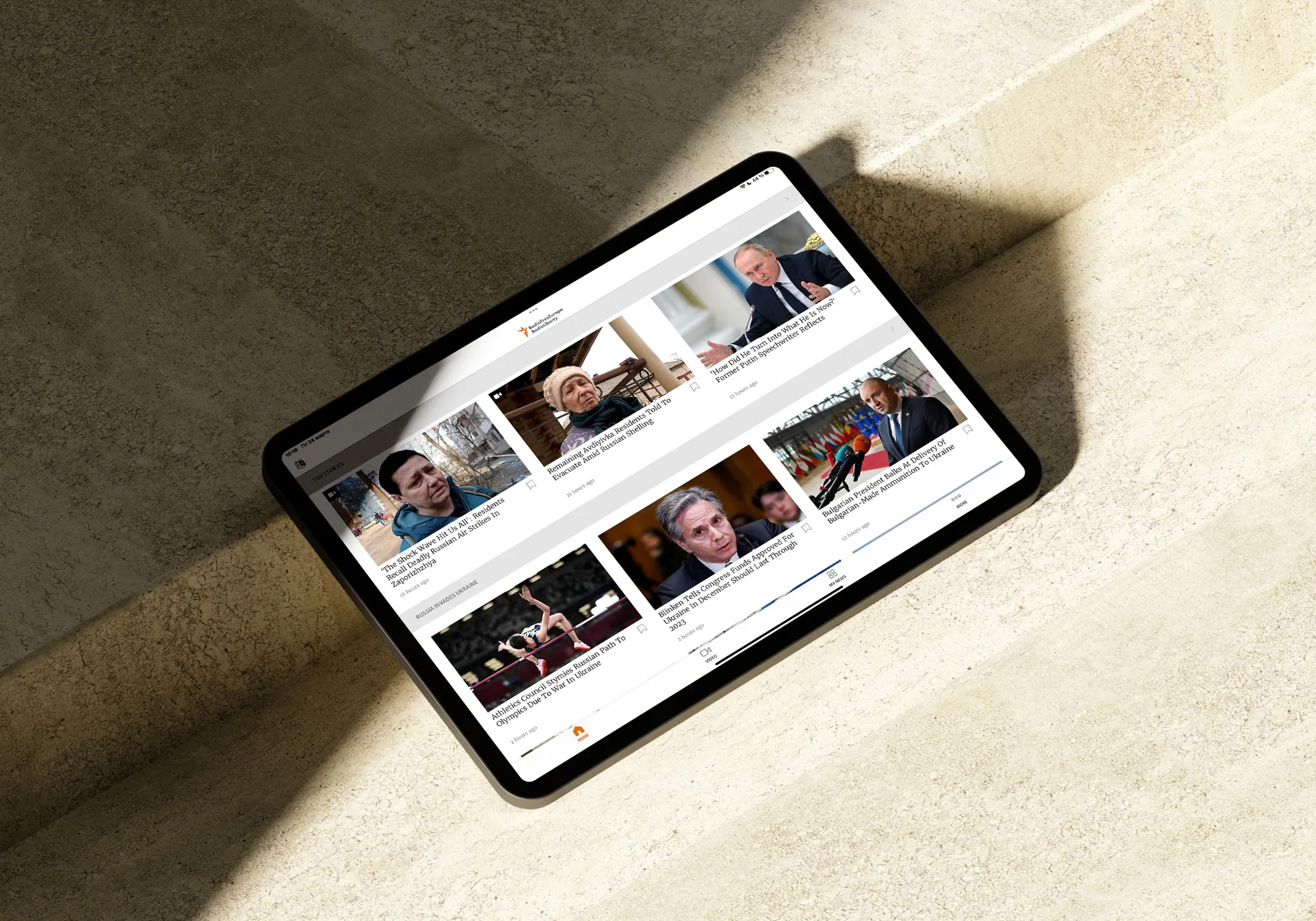 A tablet on a sunlit stone surface showing the app's top-stories grid, with the shadow of a window frame falling across it.