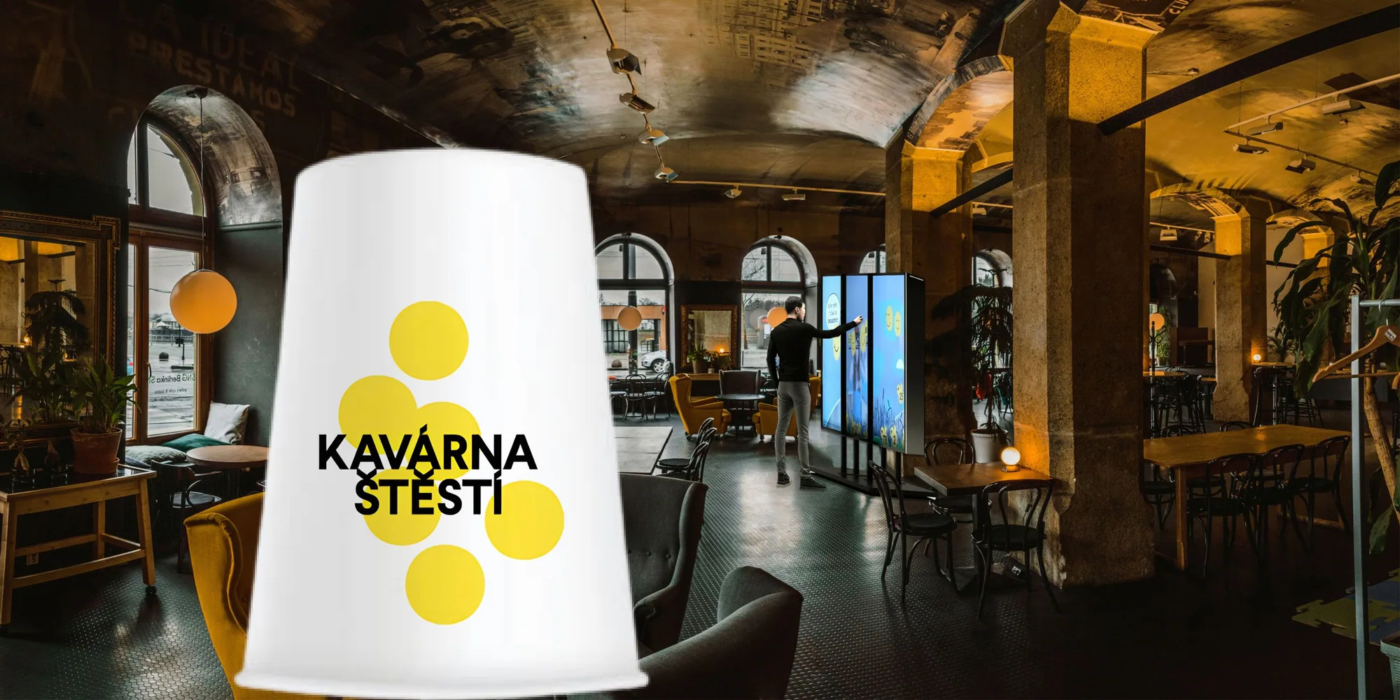 A café in a vaulted brick hall, a giant white coffee cup branded “Kavárna štěstí” standing in the foreground and a visitor playing at a trifold screen installation behind it.