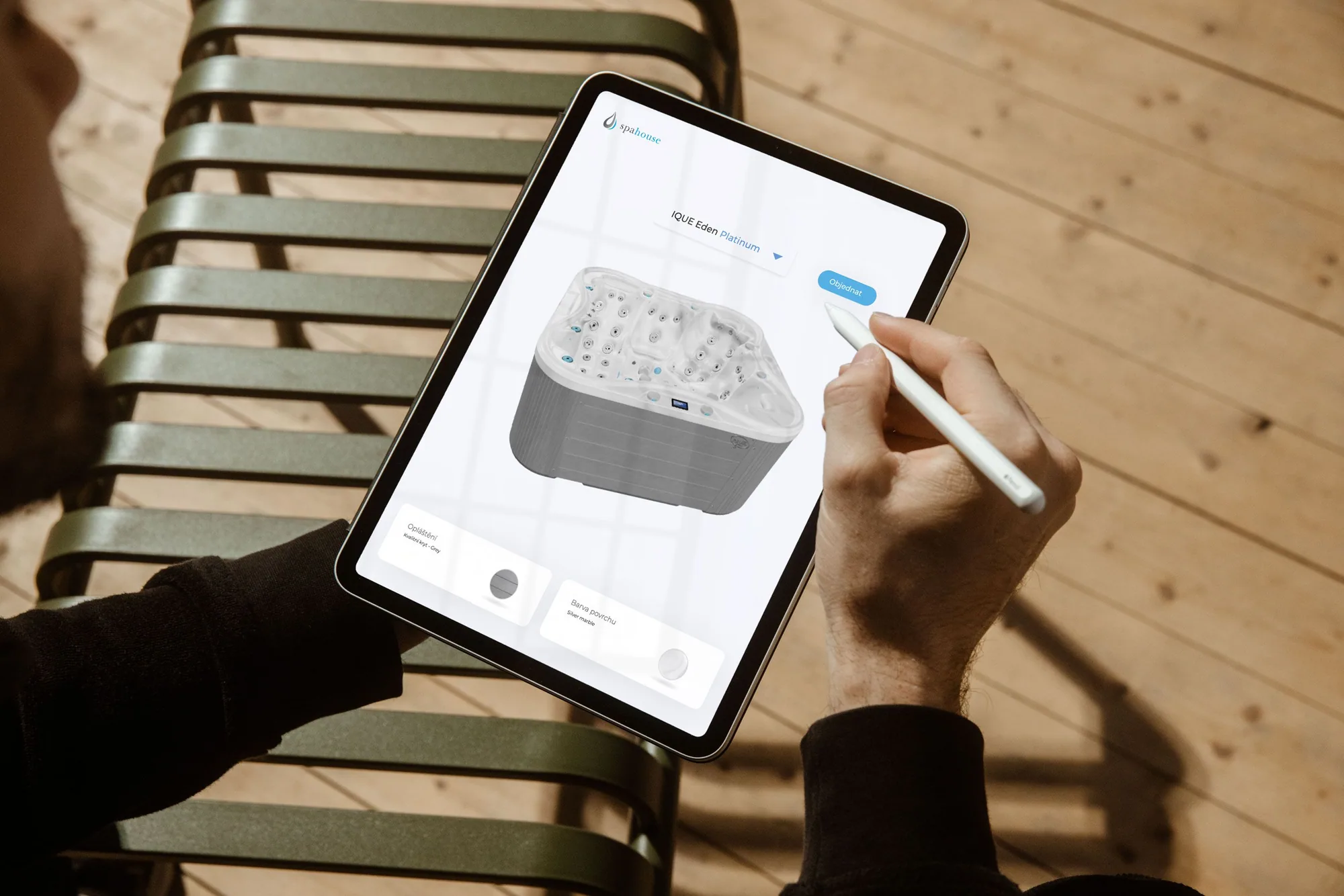 Ruka drží Apple Pencil nad iPadem s 3D konfigurátorem Spahouse: vyrenderovaný model vířivky s volbami obkladu a barvy povrchu a tlačítkem objednávky, položený na zahradním lehátku.